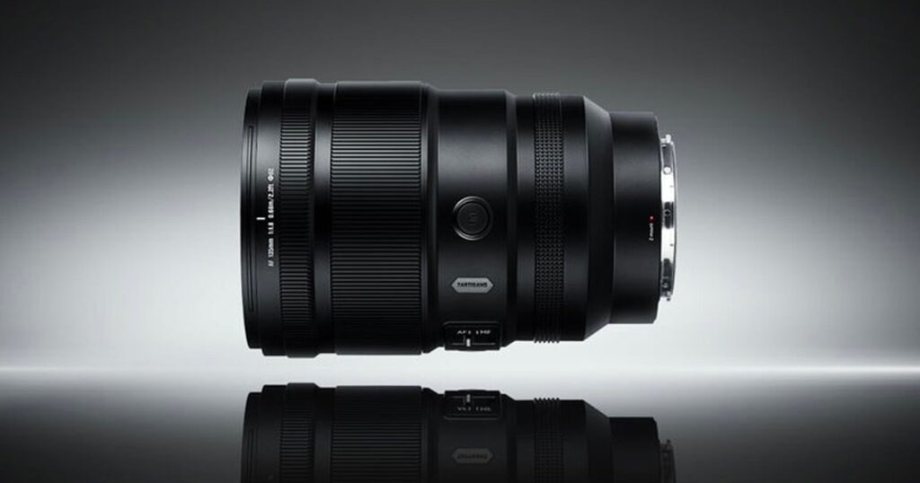 7Artisans Launches the Z-Mount’s Most Affordable 135mm f/1.8 Prime A black camera lens is positioned horizontally on a reflective surface against a gradient gray background, displaying its details and metallic lens mount.