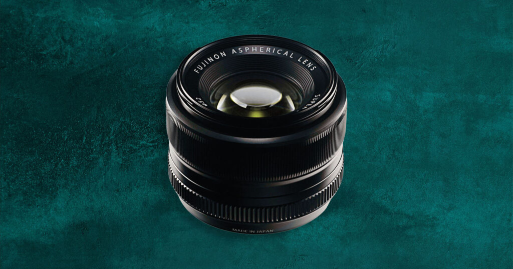 Fujifilm’s Old XF 35mm f/1.4 Is a Best-Selling Prime Lens By a Huge Margin A Fujinon aspherical camera lens is positioned upright on a textured teal background, with "Fujinon Aspherical Lens" and "Made in Japan" visible on the lens barrel.