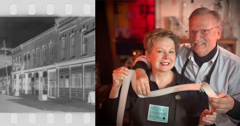 Husband and Wife are the Only People in The World Producing Film in Their Basement A grayscale negative image of a row of buildings is on the left. On the right, two smiling people hold a strip of photographic film in a studio, one wearing a "Scully Osterman Studio" name tag.