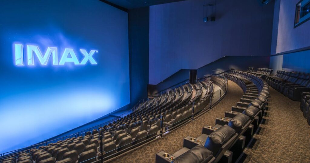 IMAX Stock Soars as Takeover Talks Revealed A modern IMAX theater with a large, curved screen displaying the IMAX logo, surrounded by rows of empty, plush black seats with cup holders. The room is dimly lit, creating a cinematic atmosphere.