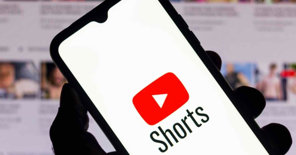 Short-Form Video Viewing on TV Surges as YouTube Shorts Hit 2 Billion Monthly Hours A hand holds a smartphone displaying the YouTube logo with the word "Shorts" below it. The background shows a blurred screen with thumbnails of multiple videos.