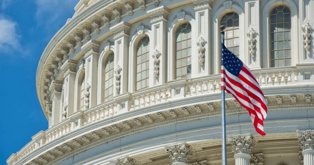 The FCC Is Hearing Public Appeals to Its Decision to Ban Foreign Drones A close-up view of the United States Capitol building’s ornate facade with arched windows, and an American flag waving in the foreground against a blue sky.