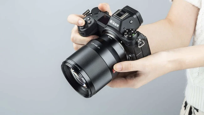 Why the 85mm f/1.8 Beats the 85mm f/1.4 for 95% of Photographers Hands holding a mirrorless camera with attached telephoto lens against gray background