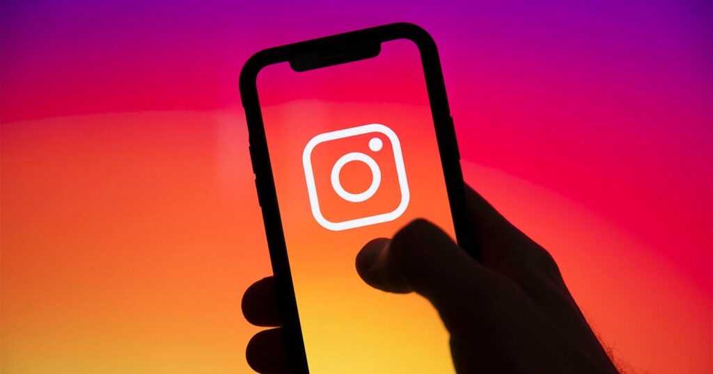 Facebook and Instagram Were Down for Nearly Three Hours Today A hand holds a smartphone displaying the white Instagram logo on a screen with a vibrant pink, orange, and yellow gradient background.