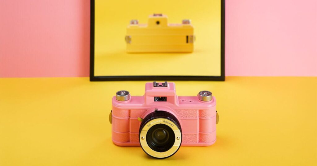 A pink vintage camera sits on a yellow surface with a pink background. Behind it, a mirror reflects the camera, but the reflection appears as a yellow camera instead of pink.