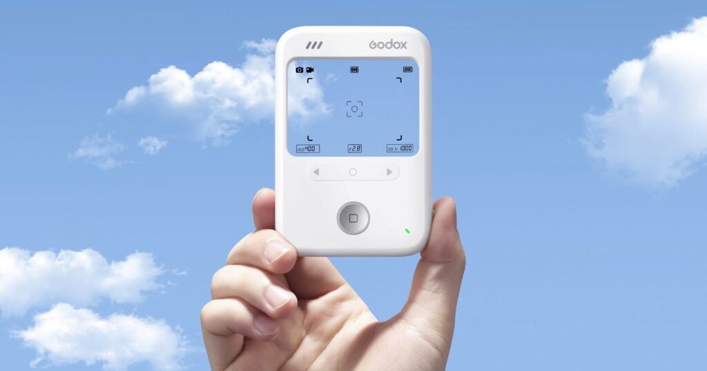 The Godox C100 is a Screenless Camera with a Transparent Display A hand holds up a white Godox electronic device with a screen displaying camera settings, against a blue sky with scattered white clouds.