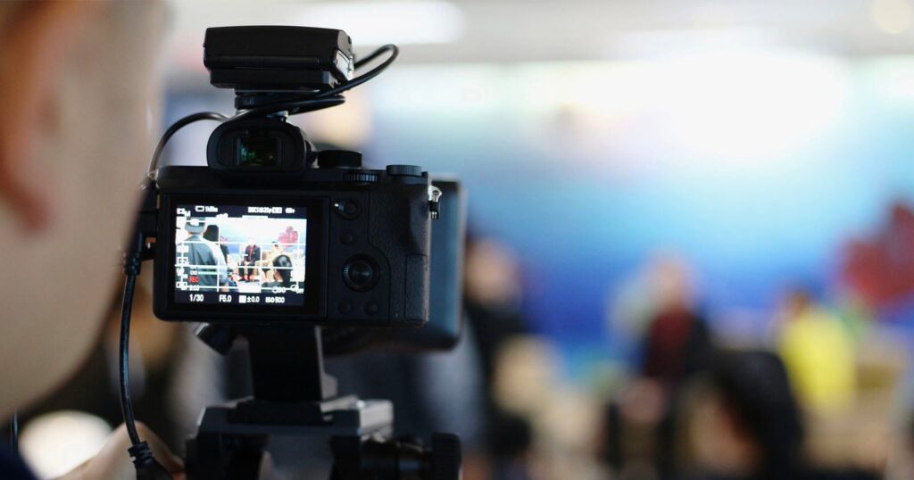 First Amendment Auditors are Taking Their Right to Film in Public to the Extreme A close-up of a digital camera on a tripod filming a group of people in a brightly lit, blurred background setting, possibly during an interview or discussion.