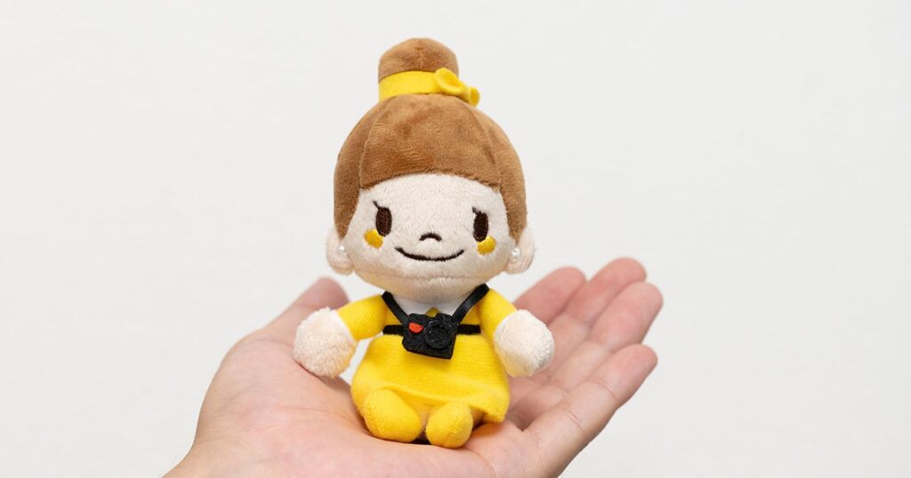 Nikon is Selling an Adorable Plushie Keychain of its Mascot A small, plush doll with brown hair in a bun and a yellow dress, wearing a miniature black camera around its neck, sits in an open palm.