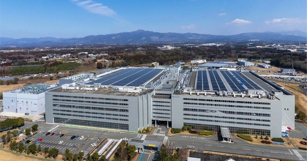 Sony’s Sensor Factory Is Back to Full Operation After Major Earthquake An aerial view of a large, multi-story Sony industrial facility with solar panels on the roof and a parking lot in the foreground.
