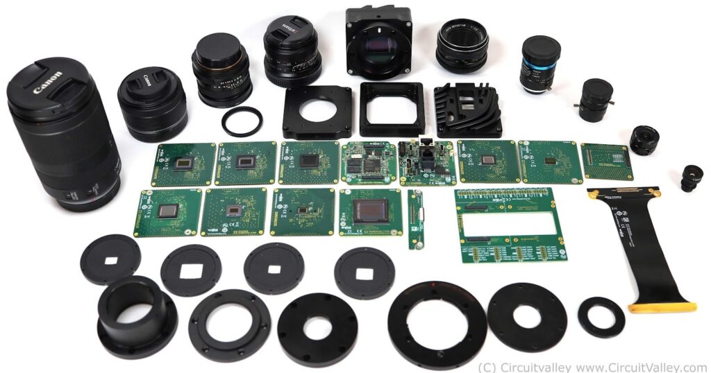 This New Open-Source Camera Has Swappable Sensors and Lens Mounts A collection of disassembled camera components, including lenses, circuit boards, sensor modules, and various mounting rings, laid out on a white surface.