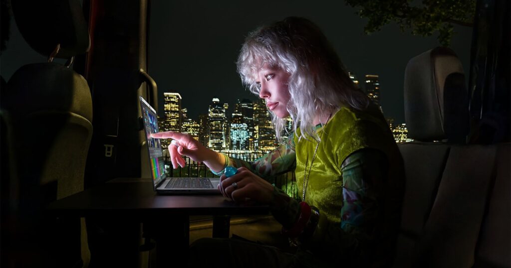 Apple Unveils Its First-Ever MacBook Pro With a Touchscreen OLED A person with blonde hair sits in the back of a vehicle at night, using a laptop while looking at a city skyline.