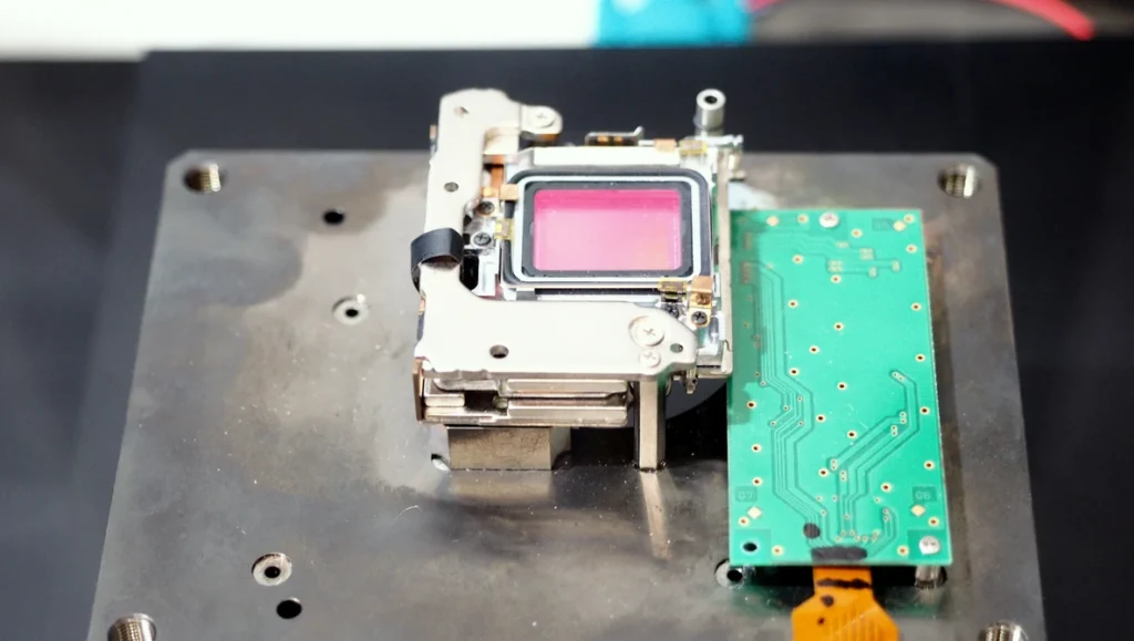 How Camera Stabilization Ratings Are Actually Measured Disassembled camera internals on a metal plate: a metal-framed module with a pink-tinted sensor beside a green circuit board