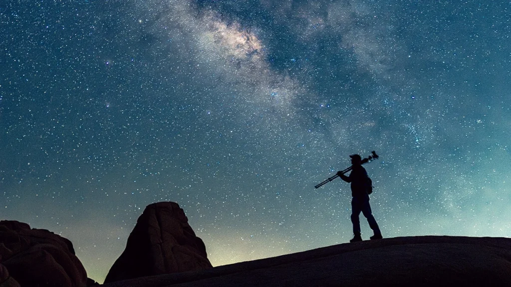 Silhouette of a person carrying a tripod over one shoulder, standing on a rock ridge beneath a bright Milky Way sky.