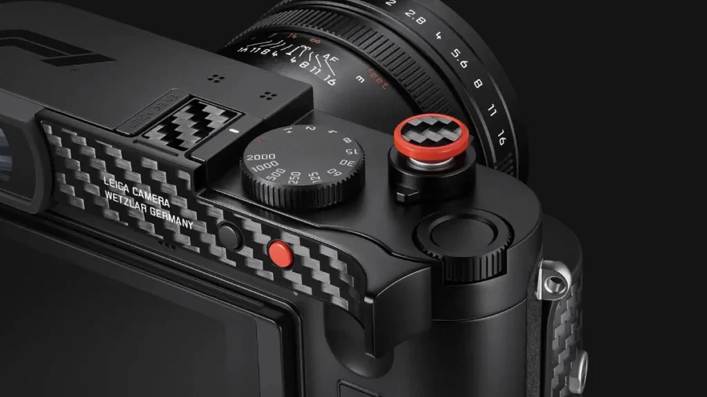 Close-up product shot of a black Leica camera's top plate and carbon-fiber grip, with a red-ringed shutter release and speed dial.