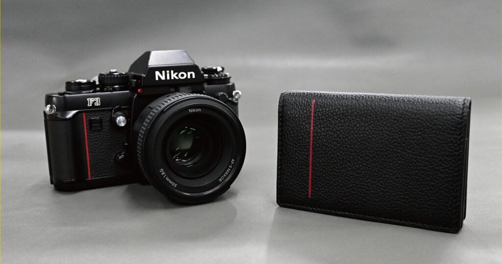 A black Nikon F3 film camera with a lens sits next to a black leather wallet featuring a vertical red stripe on a gray background.