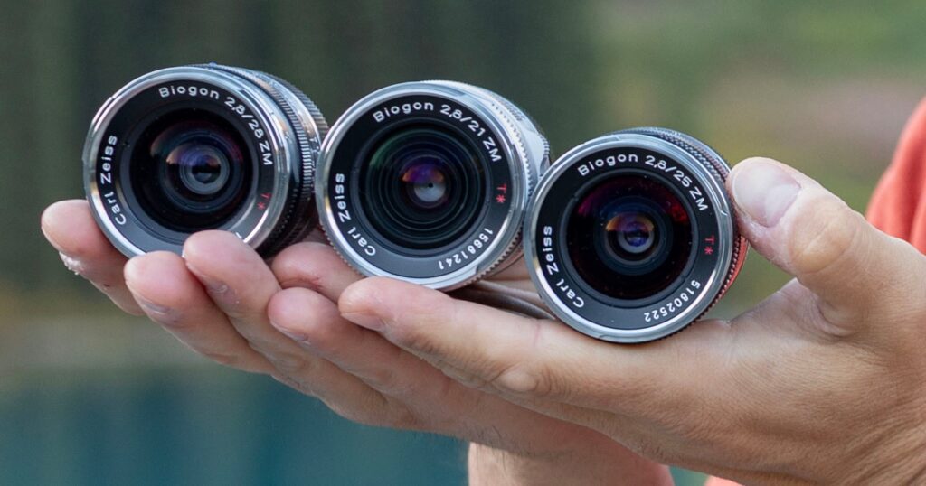 Revisiting Zeiss ZM Lenses Part 1: I’ve Always Wanted to Do This A person's hand holds three Carl Zeiss Biogon ZM lenses, labeled 2.8/28, 2.8/21, and 2.8/25, against a blurred outdoor background.