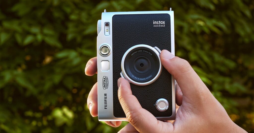 The Instax Mini Evo 2 Has a Bigger Display and Expanded Field of View A hand holds a silver and black Fujifilm Instax mini Evo2 camera against a blurred green leafy background.