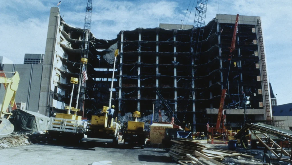 Partially collapsed multistory concrete building with exposed floors, surrounded by cranes, debris and yellow machinery.
