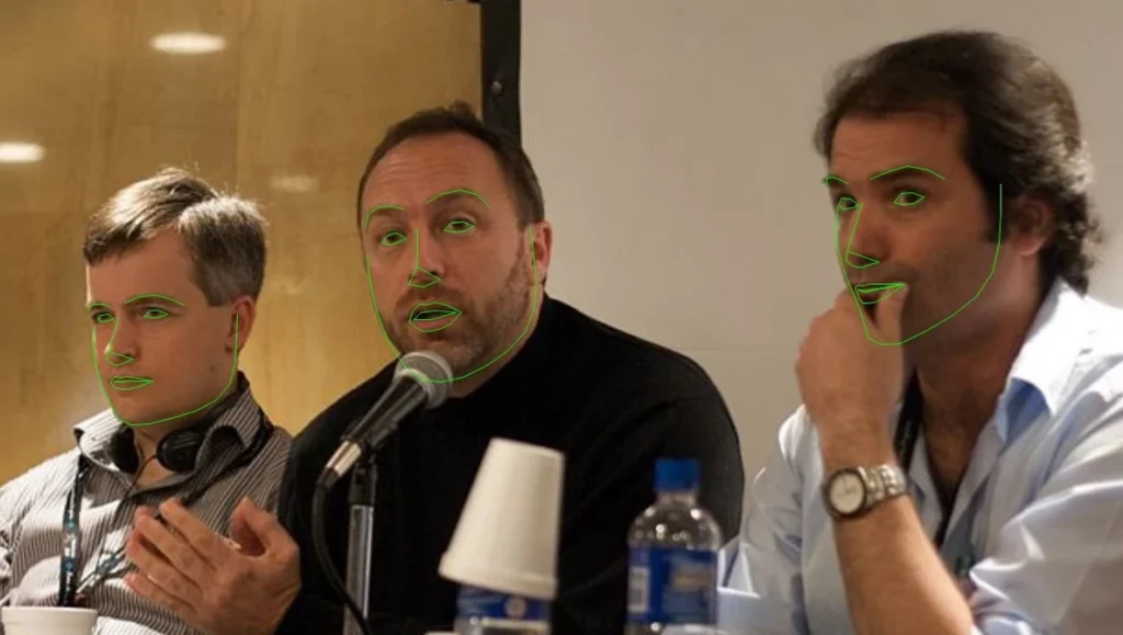 Why Passport Photos Need Strict Rules Three men on a panel, center speaker talking into a microphone, green face-tracking outlines overlaid on their faces.